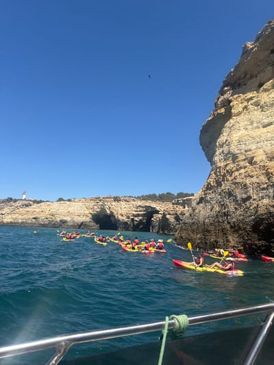 The image depicts a group of kayakers in the ocean off the coast of Lagoa e Carvoeiro, Portugal. The kayakers, wearing life vests and hats, are paddling in a cluster of yellow and red kayaks. They are positioned in front of a large, rocky cliff face with caves. In the distance, a lighthouse can be seen on the headland. The water is blue and appears choppy. The sky is clear and blue. The foreground shows a metal railing from a boat, suggesting the photo was taken from a larger vessel.