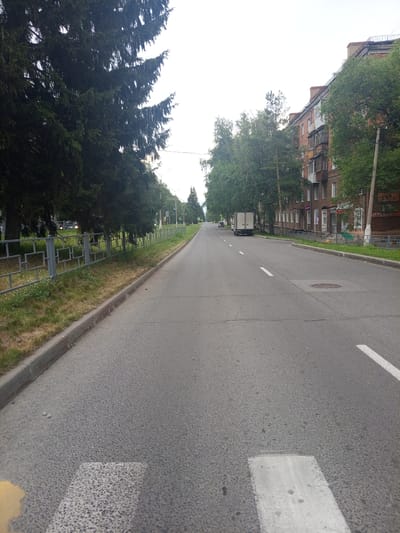 This image depicts a street scene in Yurga, Russia, likely during the daytime under overcast skies. The perspective is from a low angle, looking down a paved road that curves slightly to the right.

On the left side of the road, a dense row of tall evergreen trees borders a sidewalk and a decorative metal fence with a geometric pattern. Beyond the fence, the ground is covered in grass.

On the right side of the road, a multi-story brick apartment building is visible, with balconies and windows. Trees with green foliage grow between the building and the road. Several signs are affixed to the building, including one with "АПТЕКА" (pharmacy) written in red. A utility pole stands near the building.

The road itself is asphalt, marked with white dashed lines to delineate lanes and solid white lines for a pedestrian crossing in the foreground. A white box truck is parked on the right side of the road further down. Traffic lights are visible in the distance.

The overall environment appears to be a residential or mixed-use urban area, characterized by a mix of greenery, residential buildings, and infrastructure.