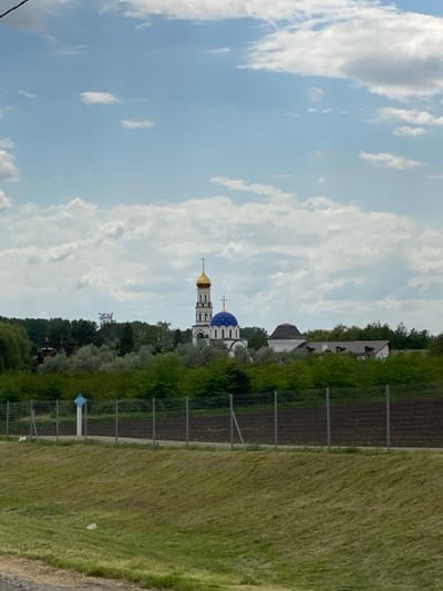 The image shows a landscape with a church in the background. The church is Orthodox with a blue and gold dome. There is a field and a fence in the foreground. The sky is cloudy. The location is Plastunovskaya, Russia.