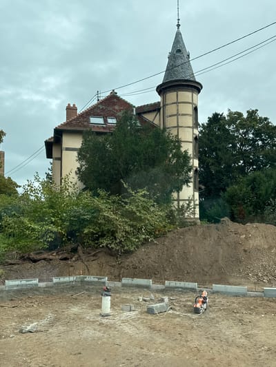This image depicts a construction site in Scherwiller, France, with a distinct building in the background under an overcast sky. In the foreground, an unpaved area of disturbed earth shows a partial, curved perimeter of light gray concrete curbing blocks laid on the ground. A white PVC or concrete standpipe is centrally located within this curved section, with several loose concrete blocks nearby. To the right of the standpipe, an orange and white cut-off saw with a circular blade is positioned on the ground, connected by a white hose. String lines for alignment are visible along the top edge of some curbing blocks.

Behind this active construction area, a large mound of dark brown excavated soil rises, partially covered with dense green bushes and exposed tree roots on its left side. Above the dirt mound, a multi-story building features light beige walls with dark timber-frame detailing. Its steeply pitched main roof is covered with red-brown clay tiles, and two rectangular skylight windows are visible. A brick chimney stack is present on the left side of the roof, and an antenna structure is mounted on the ridge.

A prominent, circular or octagonal tower with a conical dark gray slate roof extends from the right side of the building. The tower features a similar light beige facade with dark timber-framing and several narrow, vertical openings or decorative panels. The tower is topped with a finial and a lightning rod. Lush green trees and vegetation surround and partially obscure parts of the building and tower. Several black utility lines traverse the upper portion of the frame diagonally across the sky. No individuals are visible in the scene.