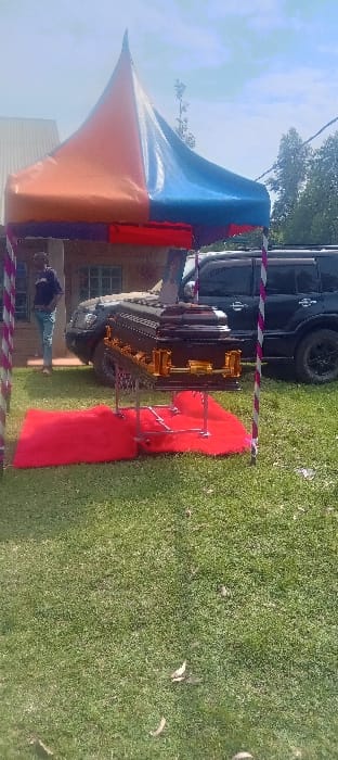 The image depicts an outdoor scene in Esise ward, Kenya, centered around funeral arrangements. In the foreground, a dark brown or black casket with ornate gold-colored hardware (handles, decorative elements) rests on a silver-colored metal folding catafalque. Beneath the catafalque and casket, a red fabric carpet or rug is laid out on the green grass.
A large, pointed canopy with sections of orange, blue, purple, and red fabric is erected over the casket, supported by four striped poles (white and reddish-purple). To the left of the canopy, a male individual stands facing right, wearing a dark t-shirt and light blue jeans, with one hand in his pocket.
In the mid-ground, behind the canopy and casket, two dark-colored SUVs are parked. The more prominent vehicle is a dark gray or black Toyota Hilux Double Cab pickup or similar large SUV. A portion of another darker vehicle is visible to its right. Behind the man and the vehicles, a light-colored building, possibly a house, with a window is partially visible. The ground is covered in green grass, with some dried leaves visible. Trees and dense foliage line the far background under a partially cloudy blue sky.