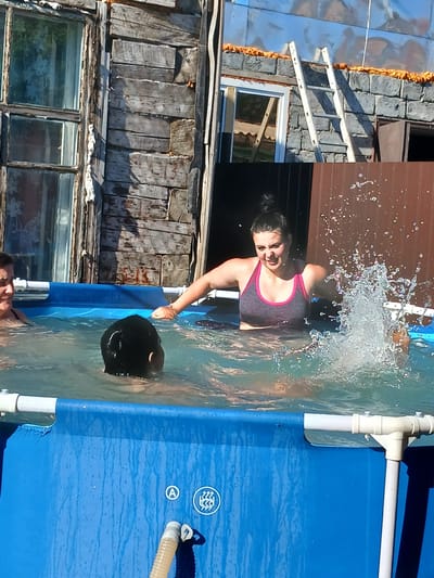 This media file shows an outdoor scene with a blue inflatable pool filled with water. Two individuals are partially visible in the pool. One person, a woman with dark hair pulled back, is in the foreground and appears to be splashing water. She is wearing a dark tank top with pink trim. Her expression suggests exertion or enjoyment. Her arm is extended, and water droplets are frozen mid-air around her, indicating movement.
Behind her, another person's head, appearing to be dark and wet, is submerged in the water, with only the top of their head visible. The pool has white PVC piping supporting its structure, and some of this piping is visible at the edge of the frame. The water is clear and reflects the surroundings, with small ripples on the surface.
The background is a cluttered outdoor environment. To the left, there is a weathered wooden structure with clapboard siding and visible nails. Above this, a section of a building with a corrugated metal roof and a white ladder leaning against a stone-like facade is visible. There is also a window with a wooden frame and glass panes reflecting light. The overall impression of the setting is rustic and possibly rural. The time of day appears to be sunny, with strong shadows cast by the structures. The weather seems warm enough for swimming. There is no visible text in the media file.