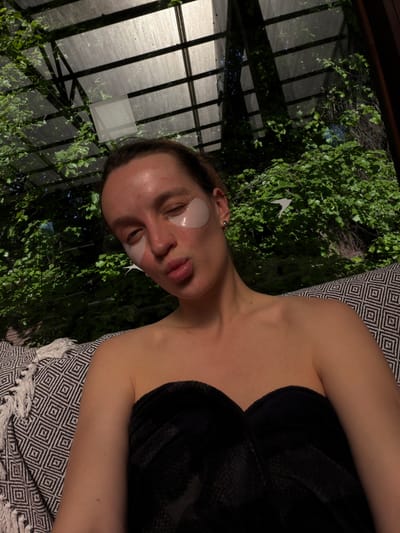 A woman is lying down in what appears to be an indoor space that resembles a greenhouse or conservatory. She has under-eye patches on her face. She is partially covered with a black towel or robe. She is lying on a patterned textile. Through the roof of the conservatory, green foliage is visible. The photograph offers no discernible information regarding the city of Tarasovka, Russia, or any details that would link the location to that city.