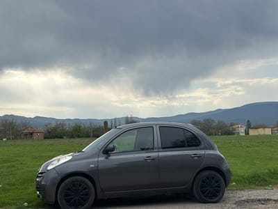 A dark grey Nissan Micra (K12 generation) is parked on a dirt path in the foreground, facing left. The car features black multi-spoke hubcaps and tinted rear windows.
Behind the vehicle, a vibrant green field stretches into the midground, indicating spring or early summer. To the left, an old, dilapidated brick house with a damaged roof stands amidst trees, some bare and some budding. Further to the right, other residential buildings, including a white house with a red roof and a yellowish building, suggest a rural village or settlement. Rolling mountains are visible in the distant background, partially veiled by the cloudy conditions.
The sky is heavily overcast with thick, grey clouds. Distinctly, streaks of virga or light rain can be seen falling from the clouds in the upper right portion of the sky. The overall lighting is dim and diffused, reflecting the gloomy, overcast weather. No people or visible text are present in the image. The scene captures a quiet, rural landscape consistent with the provided context of Padarevo, Bulgaria, under a sky threatening rain.