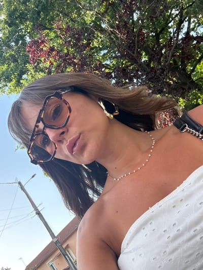 An outdoor, low-angle selfie is taken by a woman with brown hair and wearing sunglasses and a white strapless top. She is positioned in front of a blurry background of green and red foliage from a tree, with a clear blue sky visible. A utility pole with wires and a residential building with a terracotta roof are also visible in the background, suggesting an urban or suburban setting. The woman is wearing large, tortoise-shell framed sunglasses, gold hoop earrings, and a delicate pearl necklace. Her lips are slightly parted, and she appears to be looking towards the camera. The image captures a sunny day with dappled light filtering through the tree leaves. The location is identified as Savigneux, France.