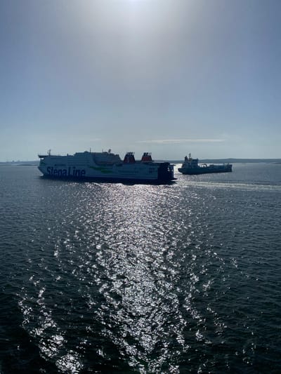 This is a vertical, medium-shot photograph of two ferries on the water under a bright, hazy sky. The ferry in the foreground is large and white, with the name "Stena Line" printed in large blue letters along its side. It also has a green and blue wave-like design. The ferry behind it is smaller and appears to be a darker color. Both ferries are oriented vertically within the frame, suggesting the photograph was taken from a higher vantage point looking down, or the image has been rotated.

The water is choppy and reflects the bright sunlight, creating a shimmering, sparkling effect across the surface. The sky is a pale blue, with some wispy clouds visible and a bright sunburst in the upper left corner, casting a bright glare. The overall impression is of a bright, sunny day, possibly in the late morning or early afternoon, given the intensity of the sunlight.

In the distance, a faint coastline or landmass is visible, suggesting the ferries are navigating a channel or near land. The presence of multiple ferries implies they are likely in a busy shipping area. The image conveys a sense of movement and travel, with the ferries sailing on the open water.

The text visible on the foreground ferry reads "Stena Line" and also "Connecting Europe for a Sustainable Future". The background ferry has some text but it is not legible.

This media file appears to be taken from the coast or from another vessel near the Asperö area of Sweden, judging by the provided location context.