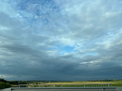 The image depicts a wide, expansive sky filled with a mixture of white and dark grey clouds, suggesting an impending storm or overcast weather. A patch of clear blue sky is visible in the upper center. The landscape below consists of agricultural fields, some appearing yellow and others green, interspersed with patches of trees. In the distance, a range of mountains can be seen, their peaks somewhat obscured by the atmospheric conditions. A metal guardrail runs horizontally across the foreground, indicating that the photograph was likely taken from a bridge or overpass. There are no people or animals visible in the scene. The image captures a rural or semi-rural environment in the vicinity of Cherna Gora, Bulgaria, characterized by agricultural land and natural topography.