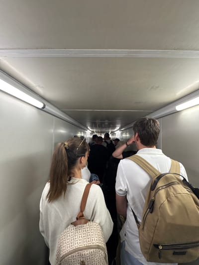 The image shows a corridor or hallway with a group of people walking through it. The corridor appears to be narrow, with white or light gray walls and ceiling. The lighting is provided by fluorescent light fixtures running along the length of the corridor. 

Several people are visible, with one man and one woman closest to the viewer, each wearing a backpack. The man is wearing a white t-shirt and the woman is wearing a white sweater. The backpacks appear to be different colors and styles. Other people can be seen further down the corridor, though their details are less clear.

The location is an aircraft jet bridge. This is inferred by the narrow passageway, the white walls, and the group of people, who appear to be passengers boarding or disembarking. It does not appear to be located in San Bartolomé, Spain.