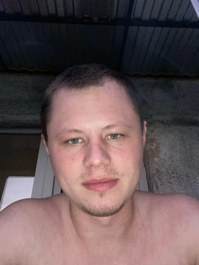 A medium shot shows a Caucasian man in his late 20s or early 30s. He has short brown hair, light green eyes, and a faint goatee and mustache. The man is bare-chested, and his skin appears flushed. He is looking directly at the camera. The background suggests an outdoor setting, possibly a balcony or patio, with a corrugated metal roof overhead and a textured wall to the right. The lighting is dim, indicating it may be evening or night. The image was taken in Beloretsk, Russia.