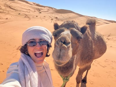 An adult female and a dromedary camel are captured in a selfie-style photograph taken in a desert environment near Rissani, Morocco. The female subject, positioned on the left, is wearing sunglasses, a light-colored beret, a light-colored turtleneck, and a matching scarf and jacket. She has dark, curly hair and is depicted with a wide, open-mouthed smile. The camel, situated to her right, faces forward with its head and part of its upper body visible; a light green rope is secured around its lower jaw. The background features extensive orange-hued sand dunes under a clear blue sky, with visible tracks and footprints on the sand surfaces.