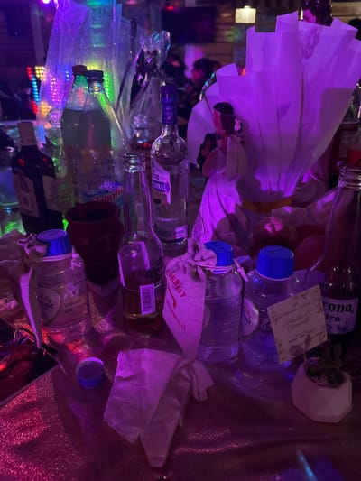 An array of objects sits under vibrant purple lighting. The scene includes several empty glass bottles of various shapes and sizes, some labeled “Corona”, “Donal”, and “Overit”. Water bottles with blue caps are also present. A small white potted plant and a card with elegant script lie among the bottles. Additional elements include a white fabric bouquet, and a brown ceramic pot. The items appear to be arranged on a reflective surface, possibly a table, adding depth to the composition.