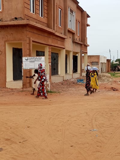 In the city of Gadao, Nigeria, a brown, two-story building with unfinished architectural features stands prominently. The building has several dark doorways and windows, some with open shutters. A sign affixed to the building reads "IJAMULSU MEDICINE STORE" and lists various services. In front of the building, a sandy dirt road is visible, with some debris scattered on the ground. Three young people are walking on the road. The person furthest to the left is wearing a white dress and is turned away from the camera. To their right, a person in a red and black patterned dress and headscarf is walking and looking to the left. Further to the right, two young people are walking, each carrying a white bundle wrapped in plastic on their heads. The person on the left is wearing a dark floral dress, and the person on the right is wearing a yellow dress. The sky above is overcast and a light grey.