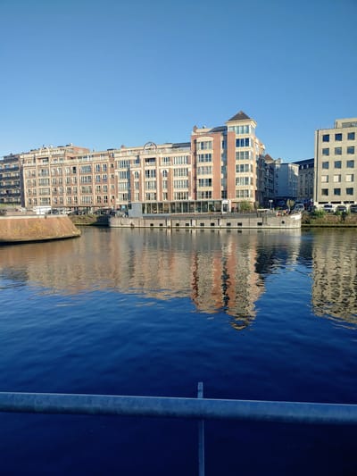 A canal runs through a city, with buildings on the far bank. A long, flat-bottomed boat is moored on the canal. The water reflects the buildings and the boat. The sky is clear and blue. In the foreground, a metal railing runs horizontally across the image. The scene appears to be in Kortrijk, Belgium.