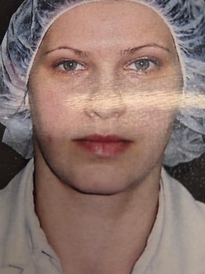 The image shows a close-up, frontal portrait of a young person wearing a white hairnet and a white garment. The subject's face is centered in the frame, with their eyes open and looking forward. They have light-colored skin, light eyebrows, and a subtle pink hue on their cheeks. Their lips are painted a reddish-pink color. The lighting in the image appears to be overhead, casting some shadows on the face and creating bright highlights on the forehead. The background is dark and appears to be a wall or curtain. There is no discernible information in the image that would allow for identification of the location in Pabradė, Lithuania.