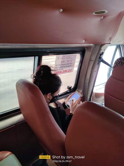 In a bus interior, a woman with her hair in a bun and wearing a black face mask is seated by the window. She is looking at her phone, which displays an image on the screen. The bus seats are upholstered in brown leather. A sign is visible on the window, containing Vietnamese text and numbers. The timestamp "2026.06.24 09:47" is present in the lower right corner, suggesting the photo was taken on this date and time. The location is specified as Ben Cau, Vietnam.