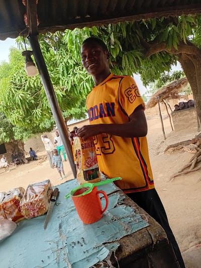 A man wearing a yellow jersey with "JORDAN 23" on it stands at a table outside a structure, likely a small shop, in Nata-Ala, Nigeria. He is smiling and holding a package of "SAWARI Buttered Bread." On the table is a red cup, a green scoop, a knife, and more packaged bread. Other people are visible in the background, standing near a brick structure and under a tree. Trees and foliage are prevalent in the surrounding environment.