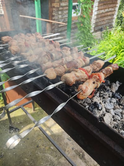 The image shows several skewers of meat being grilled over hot coals in a rusty barbecue grill. The meat appears to be pork, with some pieces interspersed with red bell pepper and onions. Smoke rises from the grill, obscuring some of the meat and the background. The grill is situated outdoors, with a wooden building and lush green foliage visible behind it. The ground beneath the grill appears to be concrete or asphalt. The overall scene suggests a summer barbecue or outdoor cooking event. The location is identified as Tavatuy, Russia.