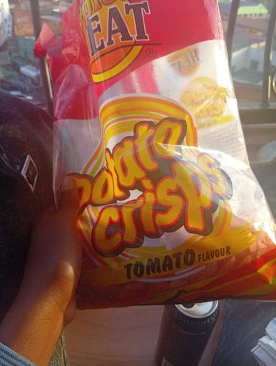 A human hand with dark skin, wearing a light blue cuff, is holding a large, clear plastic bag of "BIG EAT Potato Crisps - Tomato Flavour" in an outdoor, elevated urban setting during daylight. The bag features prominent red and yellow branding. To the lower right, a black can with a silver top, consistent with a Monster Energy drink, rests on a light brown surface. A black garment or bag with a silver buckle is visible to the left of the hand. The background reveals blurred multi-story buildings and rooftops, characteristic of the city of Murang'a, Kenya, viewed from an upper floor or balcony.