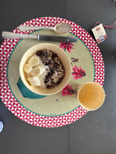 A bowl of yogurt with banana slices, chocolate shavings, and granola sits on a red and white patterned placemat, which also has a small paper cup, a knife, and a silver spoon on it. The placemat is green in the center with a stylized plant design and the words "Lira Gost" in a font. There is a partially visible piece of paper with the words "Celerina/Schlarigna" on it, placed in the upper right portion of the image. The image was likely taken at a table in Celerina/Schlarigna, Switzerland, at the coordinates 46.514 latitude, 9.859 longitude.  The bowl is blue and white with a turquoise design around the rim. The cup is white with a small black handle and appears to be empty.  The surface beneath the placemat is a dark grey, likely a table top.  The image suggests a simple breakfast meal.  The table is clean.