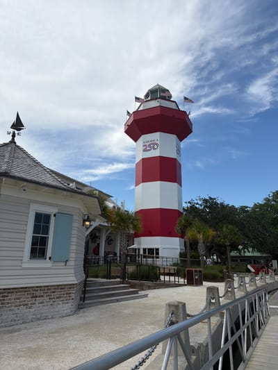 The image shows the Harbour Town Lighthouse on Hilton Head Island, South Carolina, USA. The lighthouse is a red and white striped tower with "AMERICA 250" written on its side. In the foreground, there is a white building with a blue shuttered window and a weather vane shaped like a sailboat. There are palm trees and other foliage around the lighthouse and building. A metal railing and walkway are visible along the waterfront. The sky is blue with some white clouds. A few people are visible in the background near some buildings and seating areas.