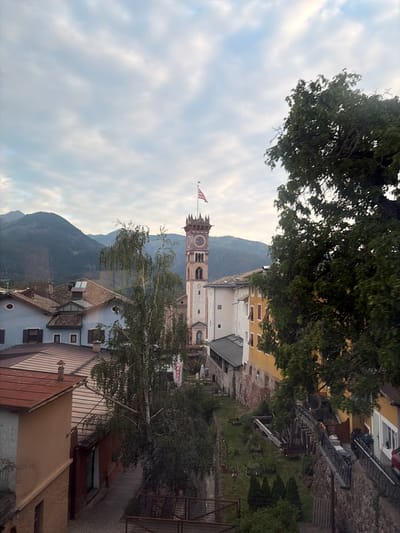 The image shows a town square in Cavalese, Italy, with a tall clock tower as the central focus. The tower is made of white stone and has a red tiled roof, with a clock face visible on its side. An American flag flies from the top of the tower. Several buildings with red tile roofs surround the square, some painted in muted colors like blue and yellow. A large green tree dominates the right side of the frame, obscuring part of the view. In the background, a range of mountains is visible under a cloudy sky. The overall scene suggests a quiet, possibly historic European town.