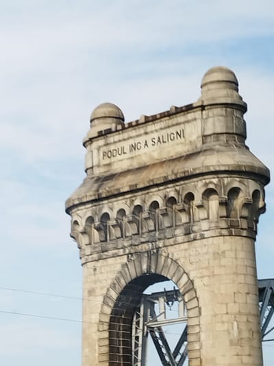 A close-up, eye-level shot captures the stone archway and a section of the upper tower of the Anghel Saligny Bridge, located in Borcea, Romania. The bridge's stonework is a pale, weathered beige, with visible mortar lines. An inscription is carved into the stone near the top of the arch, reading "PODUL ÎNCĂ A SALIGNI". The arch is ornate, featuring a row of small, semi-circular decorative arches along its upper edge. Above this, the tower structure includes stone battlements and a rounded finial. The sky in the background is a pale blue with thin, wispy clouds. In the lower right portion of the frame, a section of the bridge's metal structure is visible, hinting at the larger scale of the infrastructure. Power lines are also visible as thin, dark horizontal lines against the sky. The lighting suggests it is daytime, with the sun casting subtle shadows on the stone.