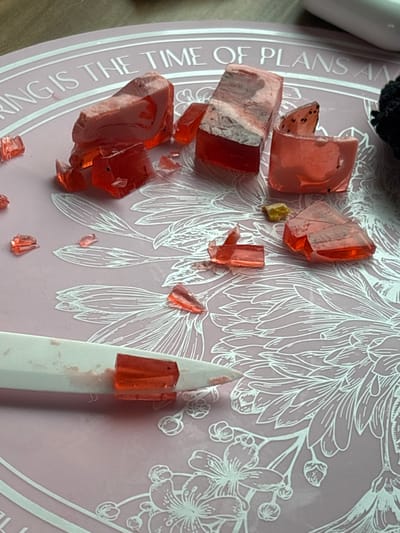 The scene, located in Barabinsk, Russia, presents a still-life arrangement on a light pink circular surface adorned with white line-art floral patterns and partially visible text reading "RING IS THE TIME OF PLANS AN". The primary subjects are multiple pieces of a translucent red, gelatinous or soap-like material. Several larger, irregularly shaped chunks are present; two prominent pieces exhibit a distinct two-layer composition with an opaque, marbled white or light grey substance layered atop the translucent red base. Numerous smaller, sharp-edged fragments of the translucent red material are scattered across the pink surface, indicating recent breakage. Small black specks are observable within some of the red pieces. In the lower foreground, a white, pointed tool, resembling a craft knife or sculpting tool, rests horizontally. Its tip and a small slot near the tip are stained with red residue and contain a small piece of the translucent red material, suggesting it was used to cut or manipulate the substance. A small, irregular, yellowish solid object lies near the center-right of the frame. A black textured object and a blurred white reflective object are partially visible in the upper right corner. No individuals are depicted.