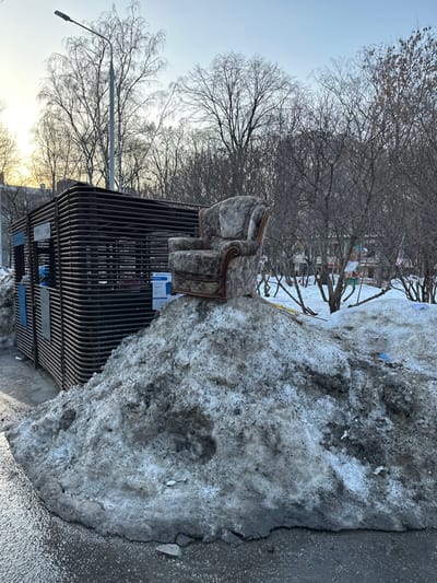 The image depicts an outdoor urban environment in Sadovaya, Russia, under clear, late-day natural lighting. A substantial, irregular mound of dirty, melting snow and ice occupies the foreground and mid-ground, showing patches of dark debris and exposed ground at its base. Centrally perched atop this snow pile is a single, worn armchair with patterned fabric upholstery in dark grey and brown tones, featuring dark wood trim along its arms and backrest. To the immediate left of the snow pile stands a dark, rectangular, cage-like utility enclosure constructed from horizontal metal slats, with several white papers or signs affixed to its left side. The ground in the immediate foreground is wet asphalt, showing cracks and small debris. In the background, numerous bare deciduous trees with thin branches are visible, suggesting winter or early spring conditions. A tall, dark streetlight pole rises on the far left, behind the metal enclosure. Faint indications of distant buildings or structures are discernible through the trees in the far background. No people are present, and no active interactions are depicted.