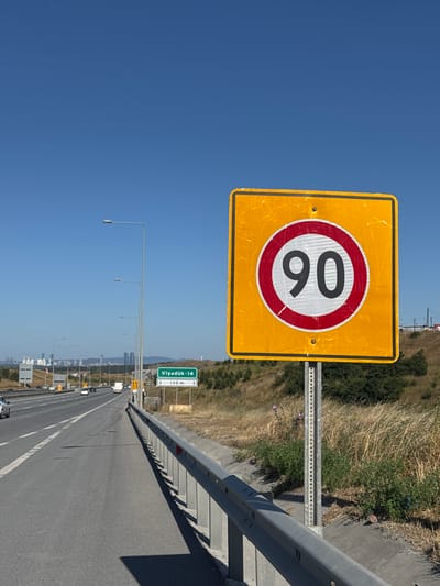 A bright yellow, square speed limit sign with a black border is prominently displayed against a clear blue sky. The sign features a circular red border with a white inner circle, containing the number "90" in black digits. The sign is mounted on a metal pole.

The scene depicts a highway, with a grey metal guardrail running along the right side of the road. The highway is asphalt and appears to be in good condition, with white lane markings visible. On the left side of the highway, there is a slope covered with dry grass and some green bushes, suggesting a rural or semi-rural setting. In the distance, buildings and possibly city structures are visible, indicating a populated area. A green road sign with white text "Viyadük-14" and "130 m" with arrows pointing forward is also visible, along with several lampposts lining the highway. Several vehicles, including cars and trucks, are on the highway, suggesting ongoing traffic. The image appears to have been taken during the daytime, with bright sunlight casting shadows. The overall impression is of a clear, sunny day on a Turkish highway.