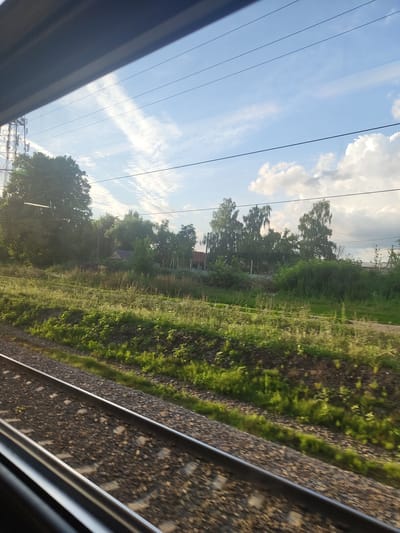 The image shows a view from a train window, looking out at a rural landscape. In the foreground, there are two train tracks. Beyond the tracks, there is a grassy field with some vegetation. In the middle ground, there are trees and a few buildings. In the background, there is a blue sky with some clouds and power lines. The scene appears to be located in the city of Isakovo, Russia, given the context provided. There are no people or animals visible in the image. The overall impression is one of a peaceful, natural environment.