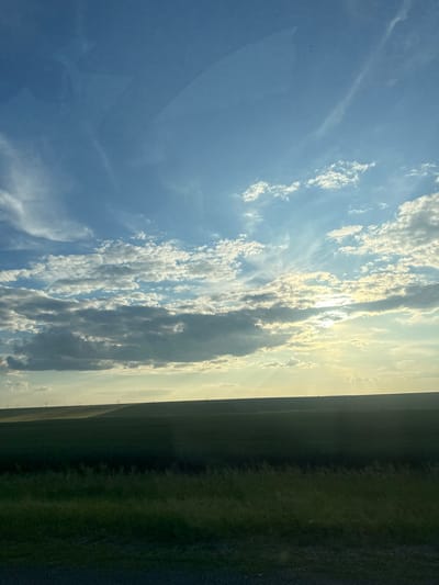 The image depicts a wide, rolling landscape under a bright, cloudy sky. The foreground is dominated by dark green grass and a patch of gravel. Behind this, a vast expanse of darker green farmland stretches across rolling hills. In the distance, silhouetted against a hazy yellow horizon, are what appear to be power line towers, indicative of rural infrastructure. The sky is a vivid blue, filled with scattered clouds, some of which are brightly illuminated by the sun, which is partially obscured behind them. Rays of sunlight pierce through gaps in the clouds, creating a dramatic effect. The overall impression is one of a serene, rural setting, likely during late afternoon or early evening due to the quality of light. There are no people or specific urban features visible, suggesting a location outside of a densely populated area.