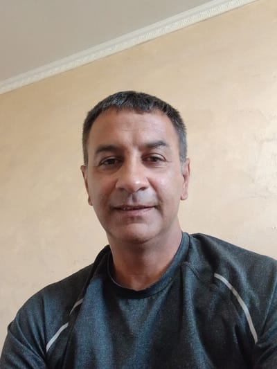 A close-up, tilted portrait shows a middle-aged man with short, dark hair graying at the temples. He has brown eyes and a slight smile. He's wearing a dark gray shirt with a light gray stripe. The background is a textured light beige wall, with a decorative white molding along the top edge. The location is specified as Topolovgrad, Bulgaria.