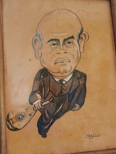 A caricature portrait depicts a bald man with large ears, dressed in a dark brown suit, white shirt, and tie. He holds a lute-like instrument. The artwork is rendered in brown tones on a textured background, framed with a simple wooden border. A signature is visible in the lower right corner in what appears to be Arabic script. The style is cartoonish with exaggerated features.