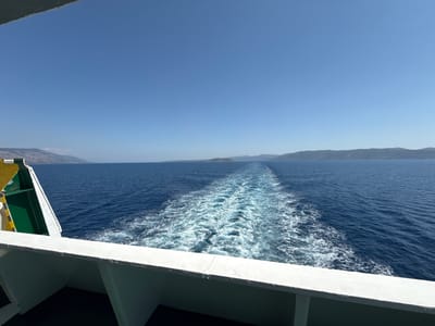 The image captures a view from the deck of a ferry boat traveling on the Adriatic Sea, likely near the town of Nerežišća, Croatia. The foreground is dominated by the white railing of the ferry, with a portion of its green and yellow hull visible on the left. The ferry is creating a prominent white wake as it moves through the deep blue water. In the distance, the hazy outlines of the island of Brač, where Nerežišća is located, can be seen along the horizon under a clear, bright blue sky. There are no people or animals visible in the frame. The scene is one of a journey at sea on a sunny day.