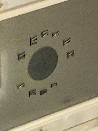 The image shows a close-up view of a metallic surface with several markings and a dark circular area in the center. The markings appear to be letters or symbols impressed into the surface. There is a distinct texture to the surface, which seems to be slightly mottled or grainy. The lighting suggests a diffused light source, casting subtle shadows. The image does not provide any context about the location, people, actions, or interactions, nor does it offer any information that could be used to ascertain its location in Krushev Dol, Bulgaria.