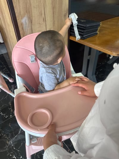 A young child is seated in a pink high chair. The child is wearing a gray t-shirt with a star pattern. The child's right hand is extended and holding a white strap attached to a stack of black menus on a wooden table. An adult's hands are visible in the foreground, with one hand resting on the high chair tray near the child's hands. The environment appears to be an indoor setting, possibly a restaurant, with dark marble flooring and wooden walls. The image was taken in the city of Ынтымак, Kyrgyzstan.