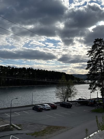 The image captures a view of a parking area adjacent to a body of water in Dom Otdykha Zolotoy Plyazh, Russia. Several cars are parked along a paved road, and a boat is also parked. A line of trees separates the parking area from the water. Power lines are visible in the sky, which is filled with clouds. The sun is shining through some areas of the clouds.