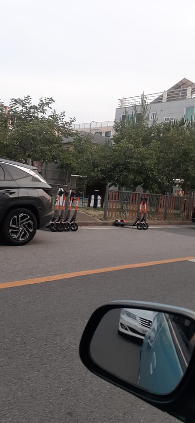 In Miryang-si, South Korea, a dark gray SUV is parked on a paved road. Adjacent to the SUV, four orange and black electric scooters are lined up upright on the sidewalk. Further down the sidewalk, two additional electric scooters are lying on their sides, one on top of the other. A wooden fence borders the sidewalk and separates it from a grassy area with trees. In the background, a multi-story building with a slanted roof is visible. The scene appears to be a street in an urban or suburban environment, with a focus on the parked vehicles and scooters. A side mirror of a car is visible in the foreground, reflecting a white car driving on the road.