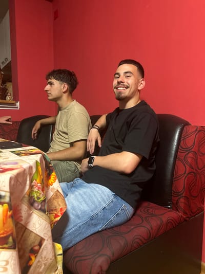 The image shows two young men seated indoors, likely in a casual home setting in Gara Elin Pelin, Bulgaria. The room features plain reddish-orange walls and a sofa or armchair upholstered in a distinctive black and red patterned fabric.
In the foreground, a young man with short dark hair and a beard is seated, smiling broadly and looking directly at the camera. He wears a black short-sleeved t-shirt, blue denim jeans, a black smartwatch on his left wrist, and a black bracelet on his right. His hands are positioned around his waist.
Behind him and slightly to his left, another young man is seated, looking downwards and away from the camera. He has curly dark hair and is dressed in a light brown or khaki short-sleeved t-shirt and light-colored pants.
A table, covered with a colorful patterned tablecloth featuring floral, fruit, and possibly autumn leaf designs, is visible in the lower right portion of the image. The general atmosphere suggests a relaxed, social gathering. There is no visible text in the image. The lighting appears even, typical of indoor artificial light or diffuse daylight.