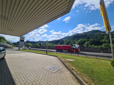 The image depicts a view from under the canopy of a gas station, likely located in Sankt Marein im Mürztal, Austria. The foreground is dominated by a paved area with interlocking concrete blocks and several metal grate covers. To the left, a silver car is parked. Beyond the paved area, a highway is visible, with a red and white semi-trailer truck driving on it. The background features a densely wooded hillside under a bright blue sky with scattered white clouds. A tall, yellow and black gas station sign with a stylized dog logo is visible to the left, and a yellow flag hangs from a pole on the right. The overall scene suggests a service area alongside a major road in a scenic, mountainous region.