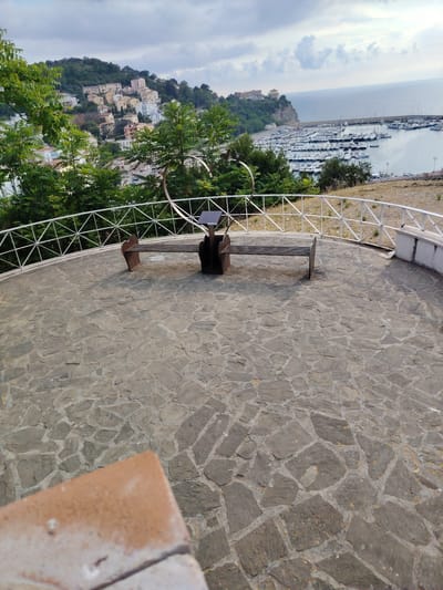 A long, flat, metal bench with a metal solar panel structure behind it sits on a circular patio paved with irregular grey stones. A white metal fence encloses the patio. In the background, a city with colorful buildings is built on a hillside, overlooking a harbor filled with sailboats and a calm blue sea under a cloudy sky. The image is taken from a high vantage point, suggesting it is part of the Belvedere di Agropoli.
