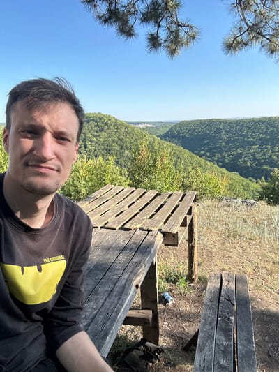A man is taking a selfie from a picnic table in a hilly, forested area with a clear blue sky above. The man is wearing a black long-sleeved shirt with a yellow design and the text "THE ORIGINAL". He is seated and looking at the camera with a neutral expression. In the foreground, there is a weathered wooden picnic table and a bench. The background features rolling green hills covered in trees, and a small settlement is visible in the distance. The overall scene suggests an outdoor recreational area. The exact location within the city of Nizhyn, Ukraine, cannot be determined from the image alone, as no specific landmarks or identifying features are visible.