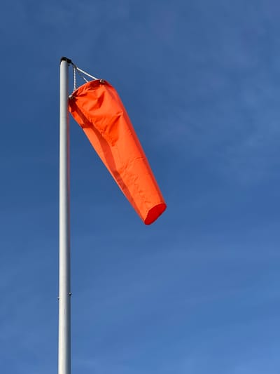 An orange windsock hangs from a metal pole against a clear blue sky. The windsock is partially inflated, indicating a gentle breeze. The pole is situated outdoors, possibly in an open area like an airfield or a coastal location, as suggested by the windsock's presence. There are no people visible in the image. The sky is cloudless and bright.