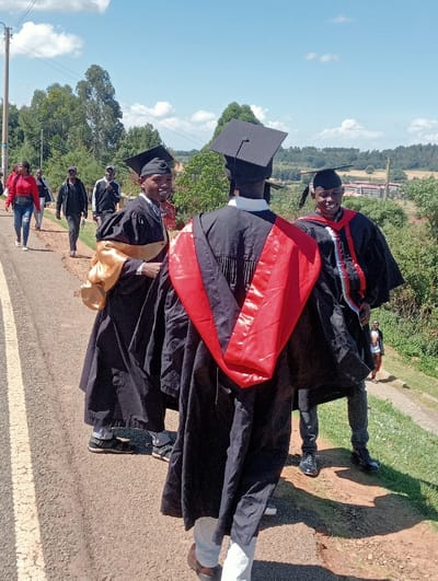 The image depicts an outdoor scene in Waseges ward, Kenya, on a clear, sunny day with scattered white clouds. Three individuals in the foreground are wearing academic graduation regalia: black gowns and mortarboards. The central figure is viewed from behind, sporting a black gown with a prominent red stole. To their immediate left, another individual in a black gown with a gold stole is seen smiling towards the viewer. To the right, a third individual, also in a black gown with a red and black stole, is smiling towards the viewer. They are positioned on the edge of an asphalt road marked with a double solid yellow line. In the mid-ground on the left, a group of approximately five casually dressed individuals, including a woman in a red jacket, are walking along the road. The background consists of a verdant landscape with numerous tall trees, and distant buildings are visible through the foliage on the right side. A dirt path runs alongside the road on the right, with one additional person discernible further back.