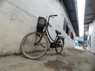 A rusty, antique bicycle with a black basket and a blue plastic-covered seat leans against a white wall. The location is a narrow alleyway in Gampongjawa, Indonesia, with a weathered concrete ground. The buildings on either side are painted white. Further down the alley, a structure with a blue roof is visible, suggesting a building or storefront where people are gathered. The sky above is overcast. The perspective is a wide angle, emphasizing the depth of the alleyway.