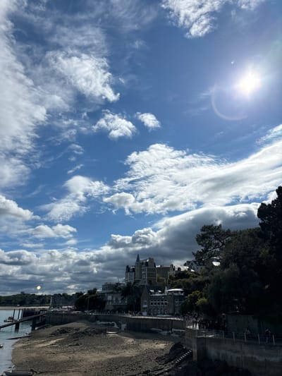 The image depicts a sunny day in Dinard, France, with a clear blue sky and scattered clouds. The sun is shining brightly, with its glare creating a halo effect. The photograph was taken from a viewpoint overlooking a low-lying, sandy shoreline. A large, white building with a pointed roof, indicative of a hotel or resort, stands on a hill overlooking the beach, with a large parking lot in front. The shore is dry and sandy, indicating that the tide is out. Numerous people are walking along the concrete path that borders the beach, while others are seen walking along the shore. Several boats are visible in the water in the distance, with a large, open-air pier leading to the shoreline. The overall scene is one of calm and relaxation, with a beautiful view of the coastline and the blue sky.