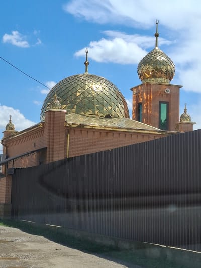 The image shows the exterior of a mosque, likely in Urus-Martan, Russia. The building features prominent golden domes with a faceted surface, reflecting the sunlight. The walls are constructed from reddish-brown brick. A tall, narrow green window is visible on one of the building's sections. The mosque is partially obscured by a dark, corrugated metal fence or wall in the foreground. The sky above is blue with scattered white clouds. A thin black wire is also visible against the sky. The lighting suggests it is daytime, possibly late morning or early afternoon, with bright sunlight casting reflections on the domes. No people are visible in the image. The scene depicts an architectural structure in an outdoor setting.