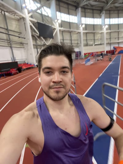 A male individual, appearing in his late 20s to early 30s, is captured in a selfie within an indoor athletic track and field facility in Tashevka, Russia. He has short, dark hair styled upwards, light skin, and a short beard. He is wearing a purple sleeveless athletic tank top with a red Nike logo on the chest, and white wireless earbuds in both ears. A black armband is visible on his left bicep. He is positioned facing the camera with a neutral expression. The background reveals a multi-lane red running track marked with white lines, and a distinct blue track section is visible to his right. Red spectator seating is arranged along the left side of the track. The facility features high ceilings with exposed white structural beams, large windows providing natural light, and supplementary artificial lighting fixtures. A large, dark rectangular display screen is mounted high on a wall in the distance, and various athletic equipment, including large blue and orange landing mats, are visible further back on the track floor.