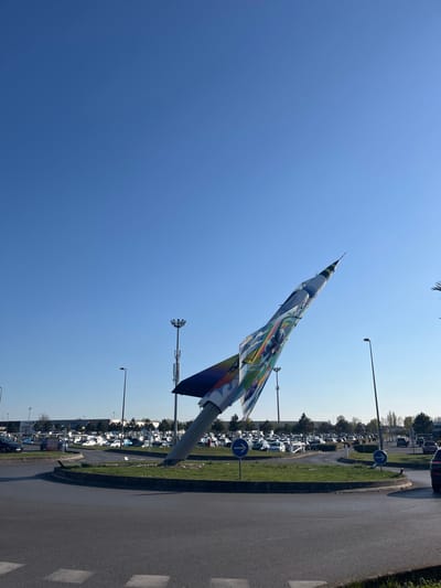 The image shows a large, colorful sculpture of a jet fighter aircraft, painted in bright colors, in a roundabout in Saint-Brice-Courcelles, France. The sculpture is positioned in a grassy area in the center of the roundabout. The roundabout is paved with gray asphalt and surrounded by a curb. There are several cars parked around the perimeter of the roundabout. The sky is a clear blue, and the sun is shining. There are some streetlights and other foliage visible in the background. The sculpture is pointing towards the right side of the image. There are two blue signs with white arrows pointing to the right.