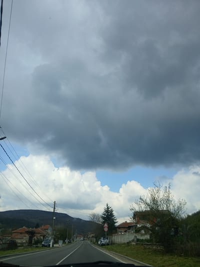 The image presents a landscape view, possibly taken from a vehicle traveling on a road in what appears to be Batovo, Bulgaria. A significant portion of the sky is covered by dark, ominous storm clouds. To the right of these clouds, there is an area of brighter, white cumulus clouds and a patch of blue sky. The road curves slightly, with houses and vegetation visible along the roadside. There's a speed limit sign with the number "30" on it. Mountains or hills rise in the background. Power lines run along the side of the road.